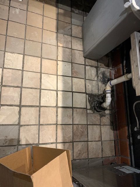 Small tiled floor with a cardboard box and a pipe opening near a wall, showing a wet or dirty corner area