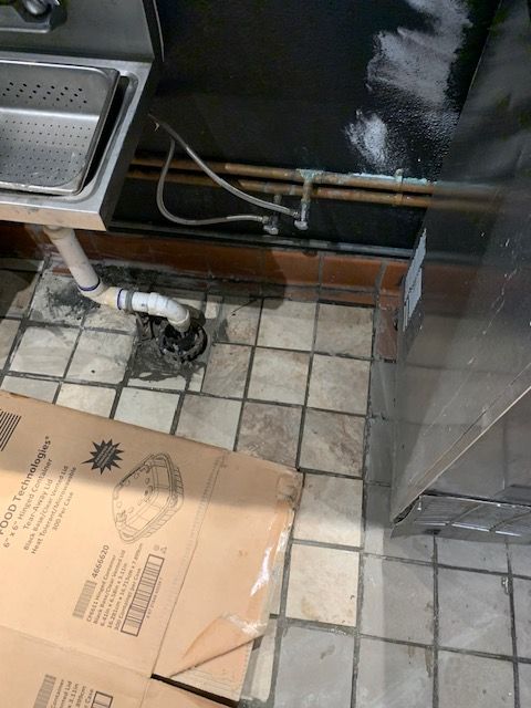 Kitchen floor with tiles, a box, and exposed pipes under a sink or counter.