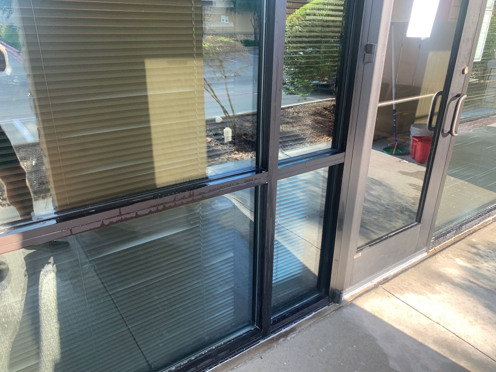 Glass storefront windows and door beside a sunlit sidewalk, with blinds visible inside.