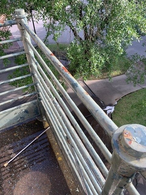 View from a balcony with rusted railings overlooking a sidewalk, grass, and trees below