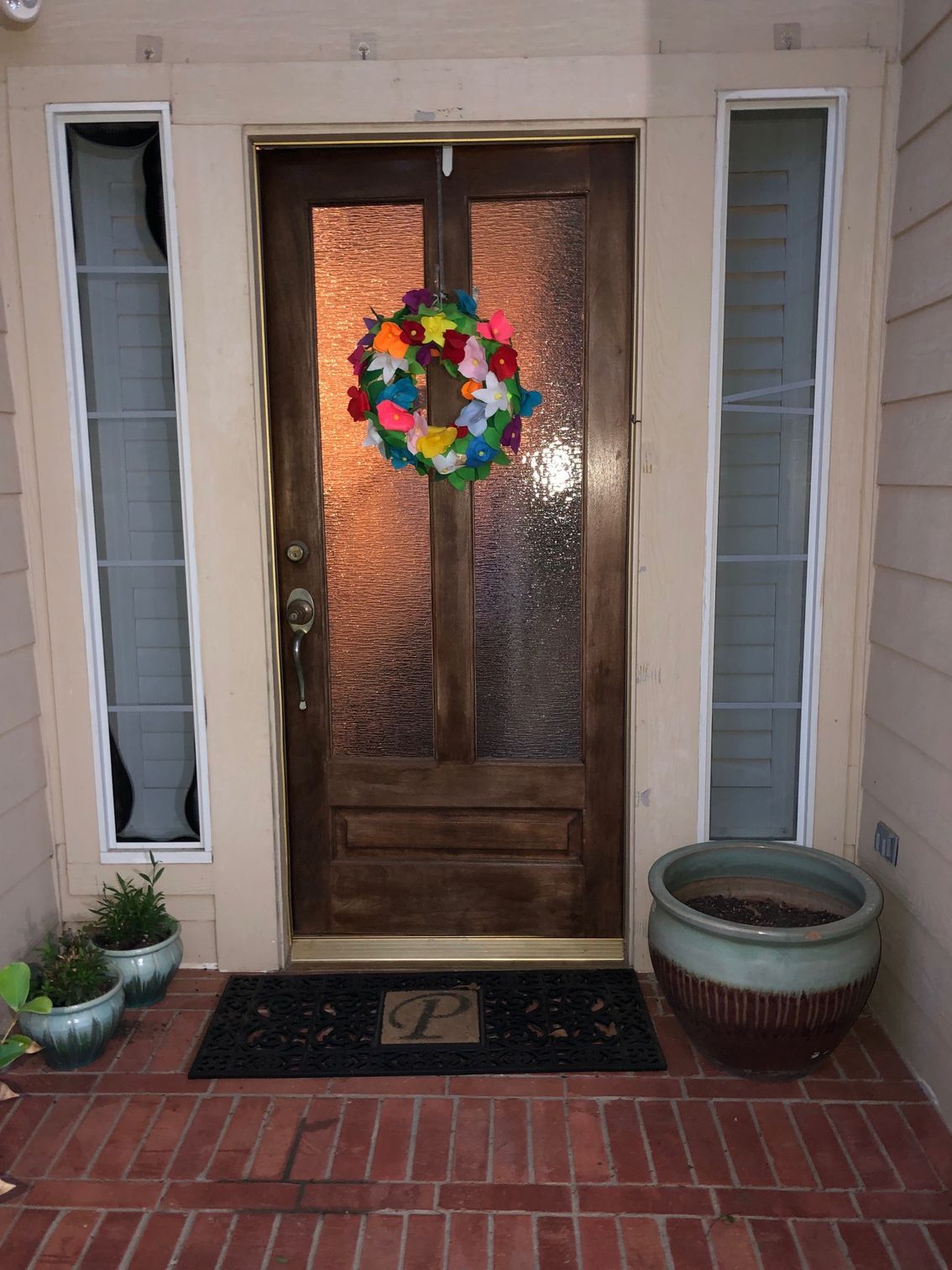 Front door with colorful wreath, flanked by tall windows, brick steps, and potted plants.