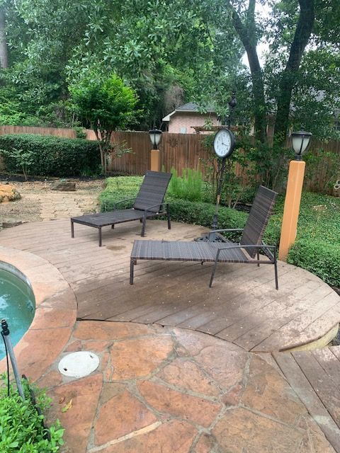 Backyard patio with two dark lounge chairs beside a pool, surrounded by greenery and a wood fence.