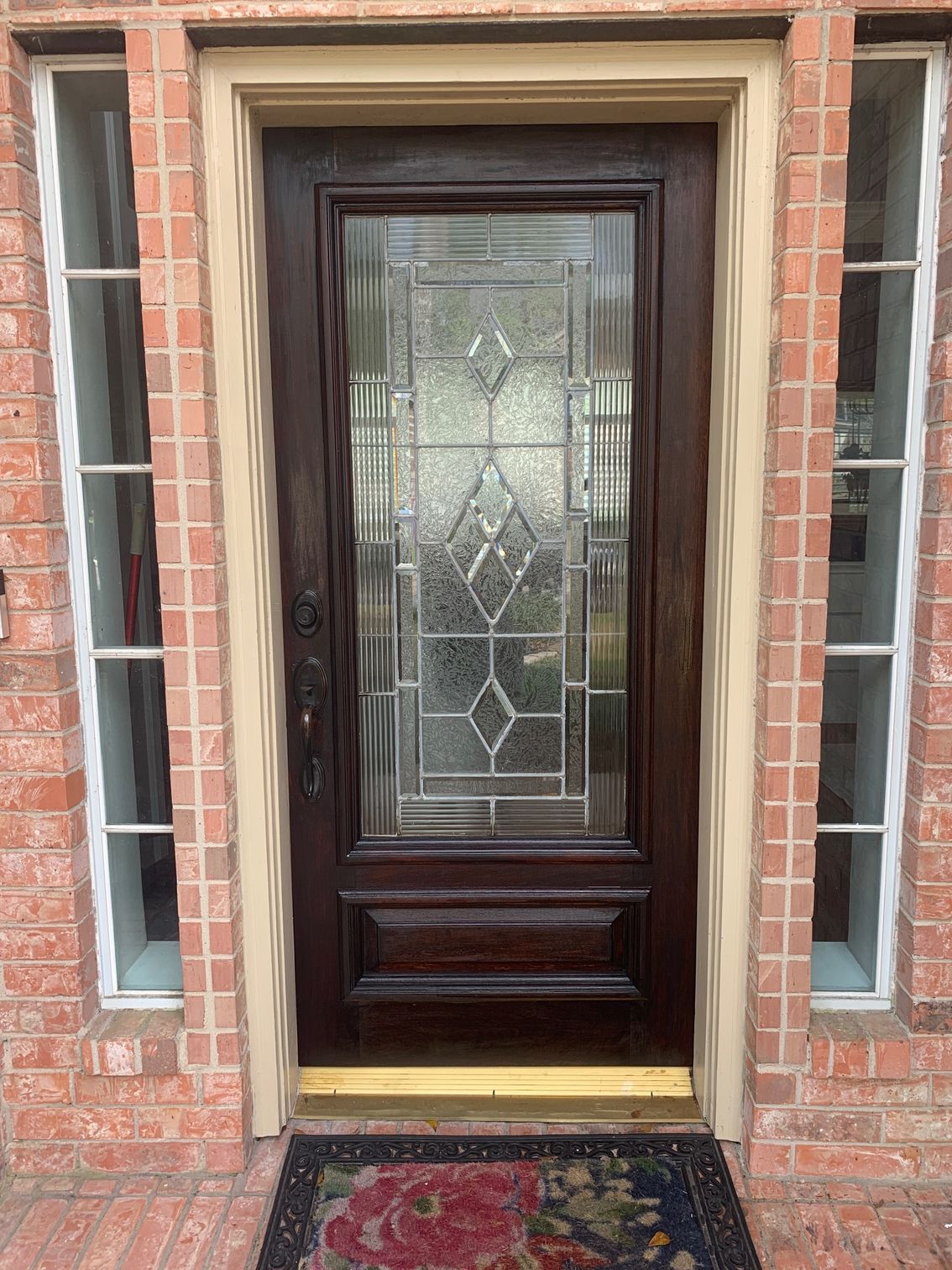 Dark wood front door with decorative glass panels and sidelights, set in a brick entryway with a doormat.