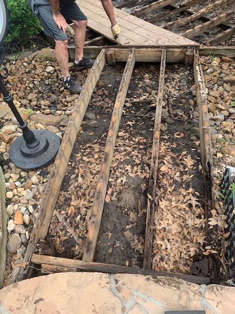 Person working on a wooden deck frame over a rocky ground, with leaves and tools nearby