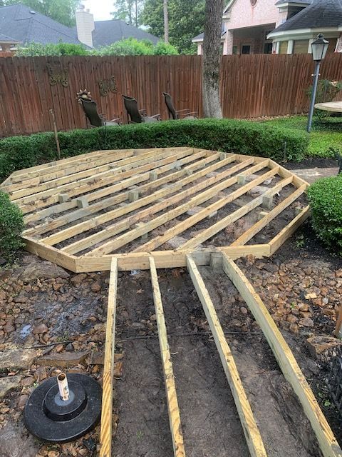 Broken wooden deck frame in a backyard, with exposed joists over a muddy hole and fence in the background
