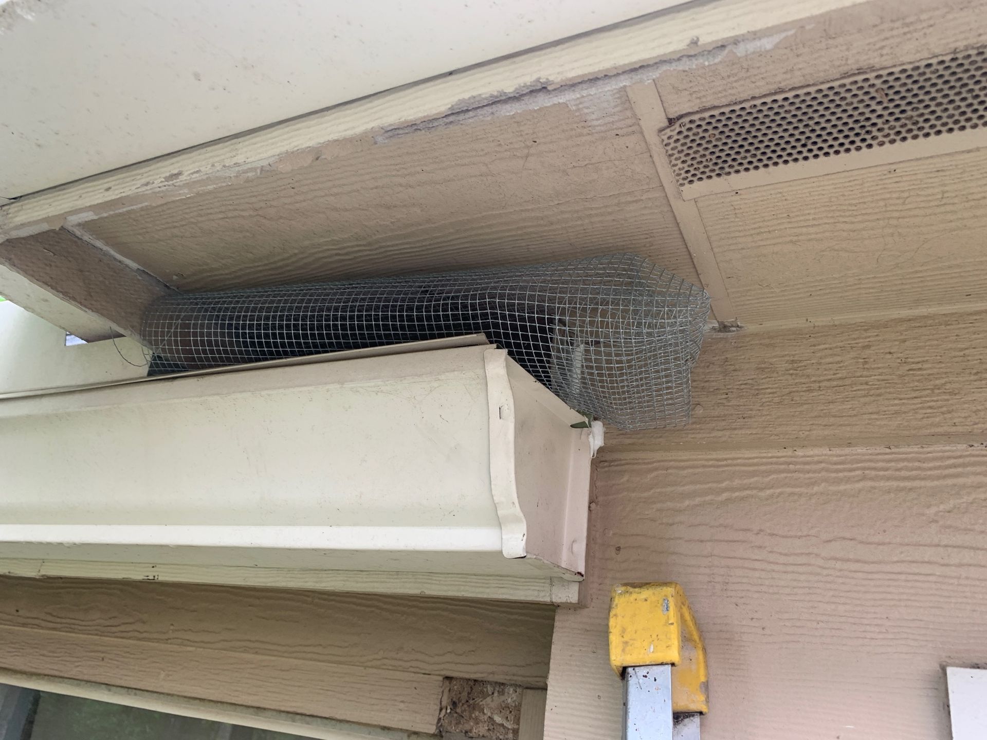 Damaged building soffit with exposed wire mesh and a yellow utility box on a tan exterior wall