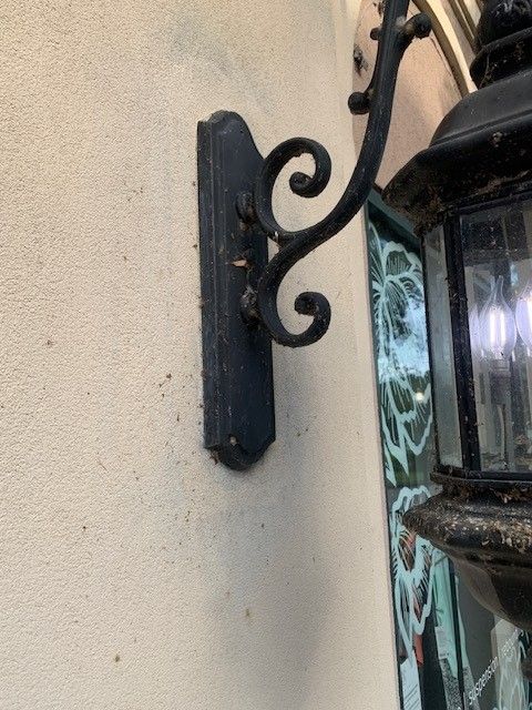 Black wall-mounted lantern bracket beside a cream stucco wall and tiled exterior trim