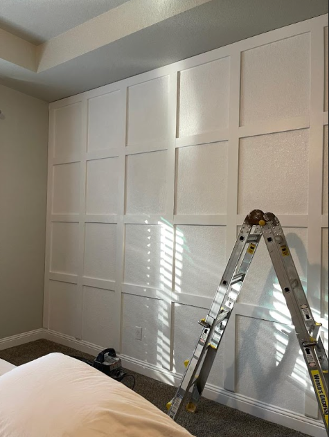 White paneled bedroom wall with a ladder leaning against it and sunlight casting window-blind shadows