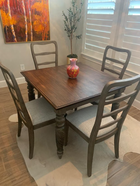 Wooden dining table with four chairs in a bright room, with a vase centerpiece near a window and wall art.