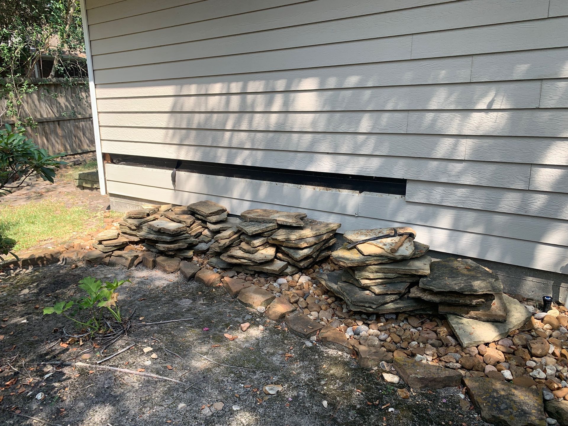 Stone retaining wall along the base of a light gray house siding, beside a dirt and gravel yard.