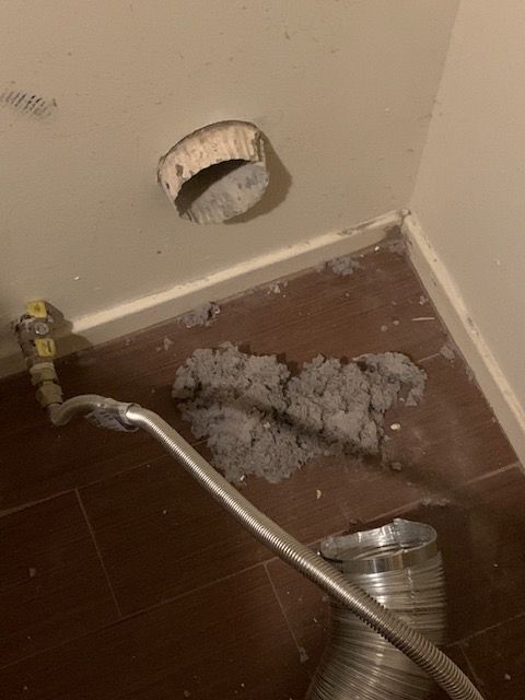 Broken wall vent with loose ducting, debris on the floor, and a detached hose in a corner.