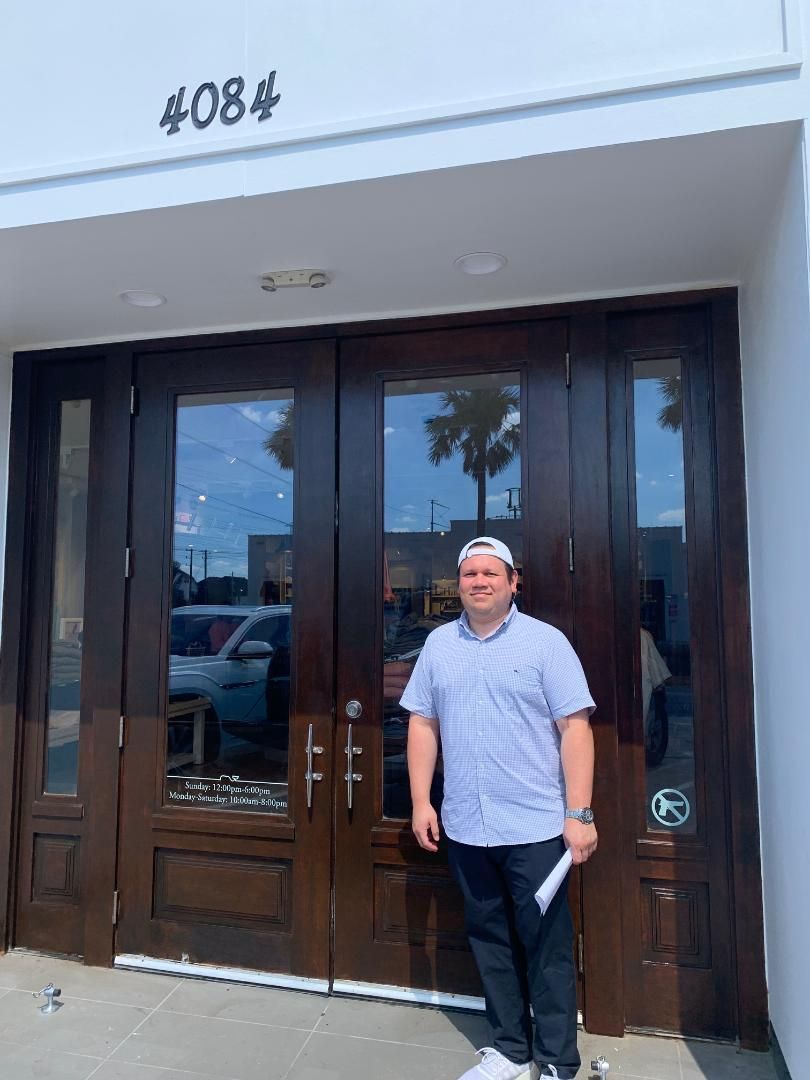 Man standing outside glass double doors marked 408, wearing a light blue shirt and dark pants.