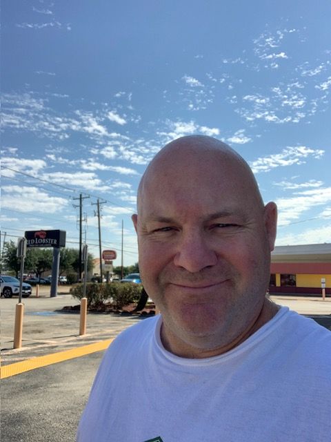 Smiling bald man in a white shirt outdoors under a blue sky with parking lot and road signs nearby