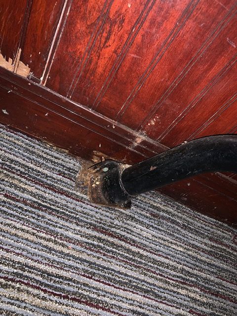 Black cable passes through a wall hole onto a striped carpet beside a red wood wall panel.