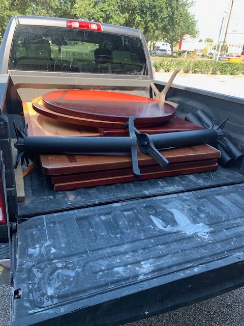 Pickup truck bed loaded with stacked wooden chairs and a round table top on a sunny driveway
