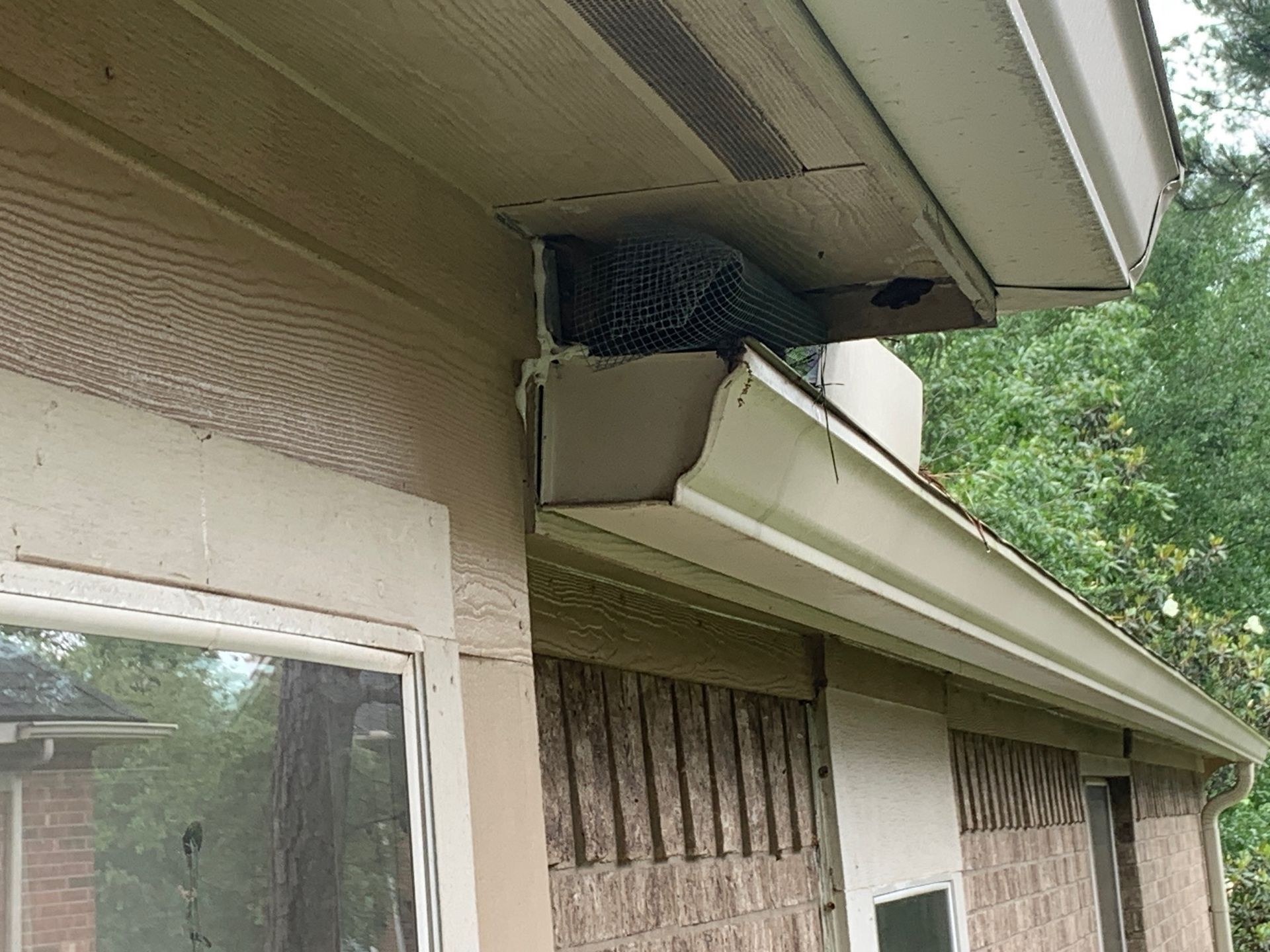 Broken soffit and hanging gutter under a house eave, with a black mesh repair patch visible.