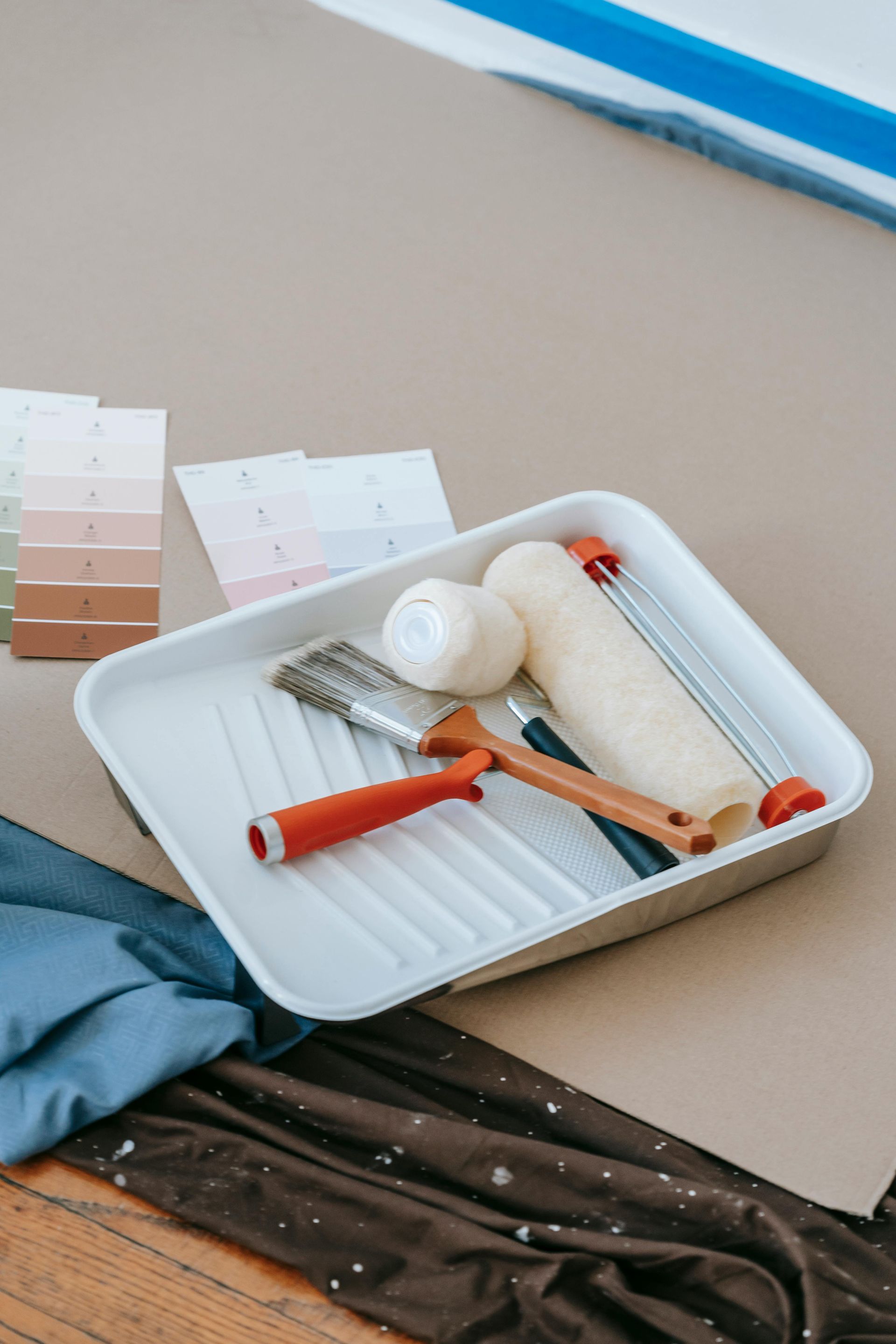 A tray with paint rollers , brushes , tape and paint samples on a wooden floor.