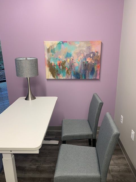 Small office corner with a white desk, gray chair, table lamp, and colorful wall art on a lavender wall.
