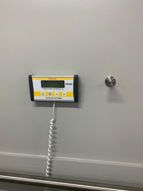 Wall-mounted electronic display with a coiled cord on a light gray wall, beside a small round fixture.