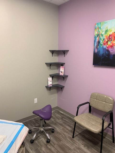 Empty exam room with mauve walls, a stool, chair, and corner shelf holding pamphlets.