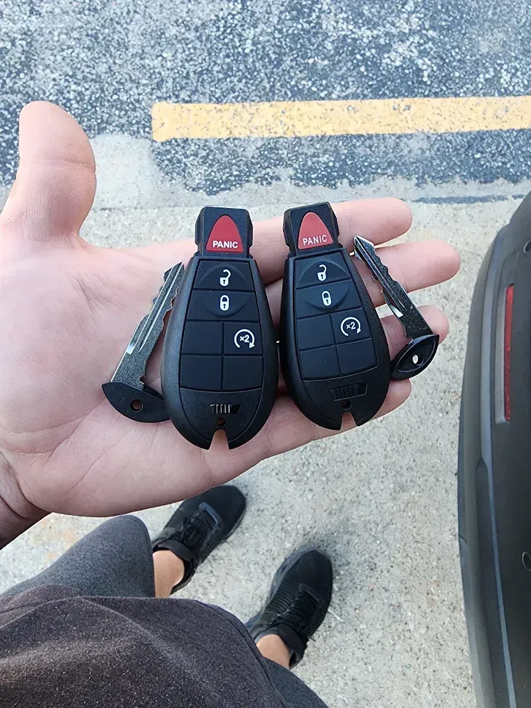 Hand holding two black car key fobs with red buttons on a parking lot surface