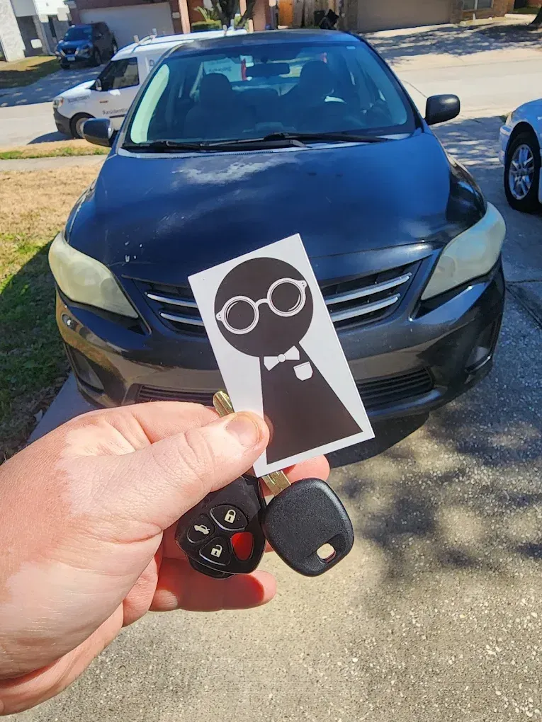 Hand holding car key fob in front of a dark blue sedan parked outdoors; cartoon face card in foreground