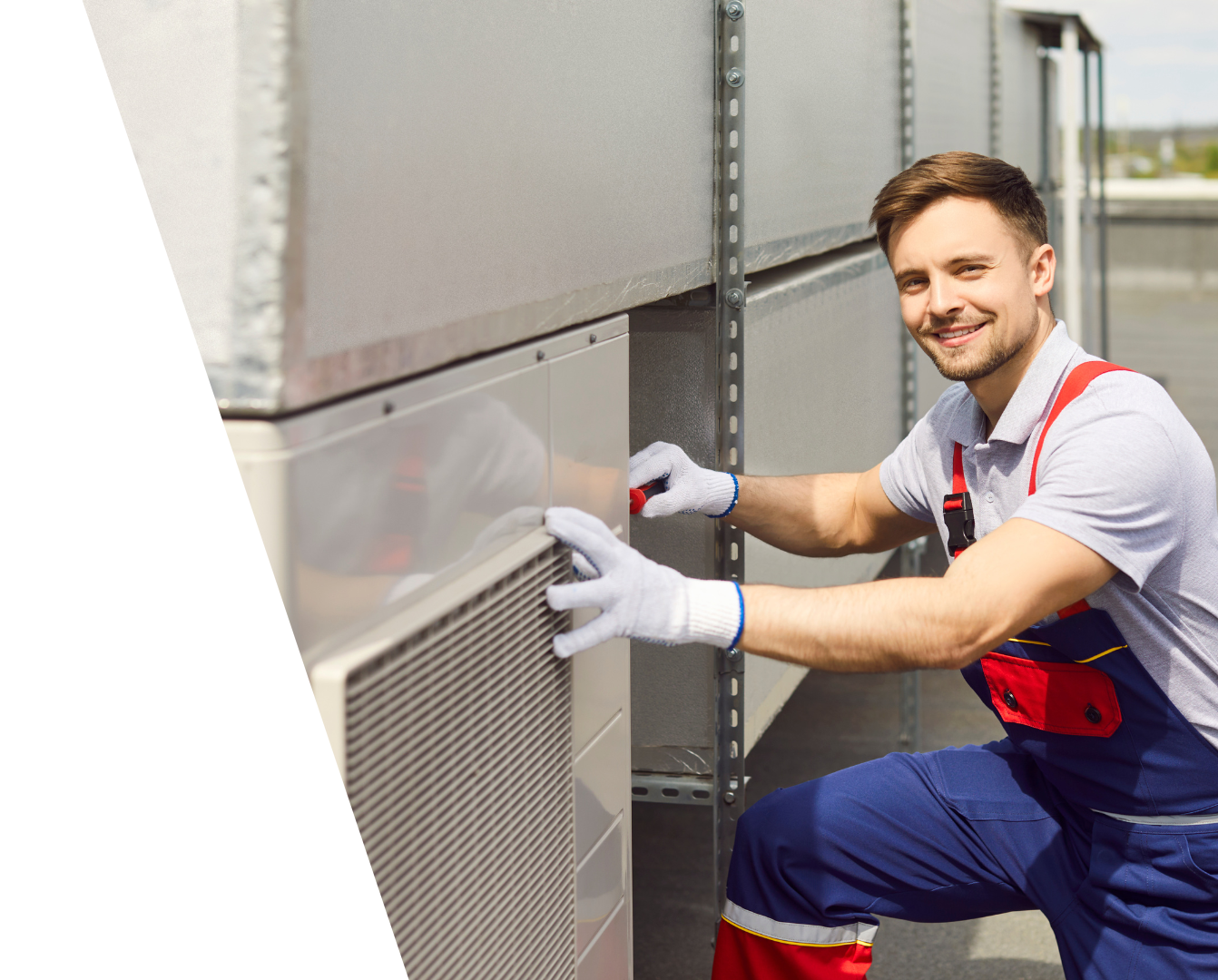 Contact Us! Technician in red overalls adjusting a metal HVAC unit outdoors, smiling at the camera