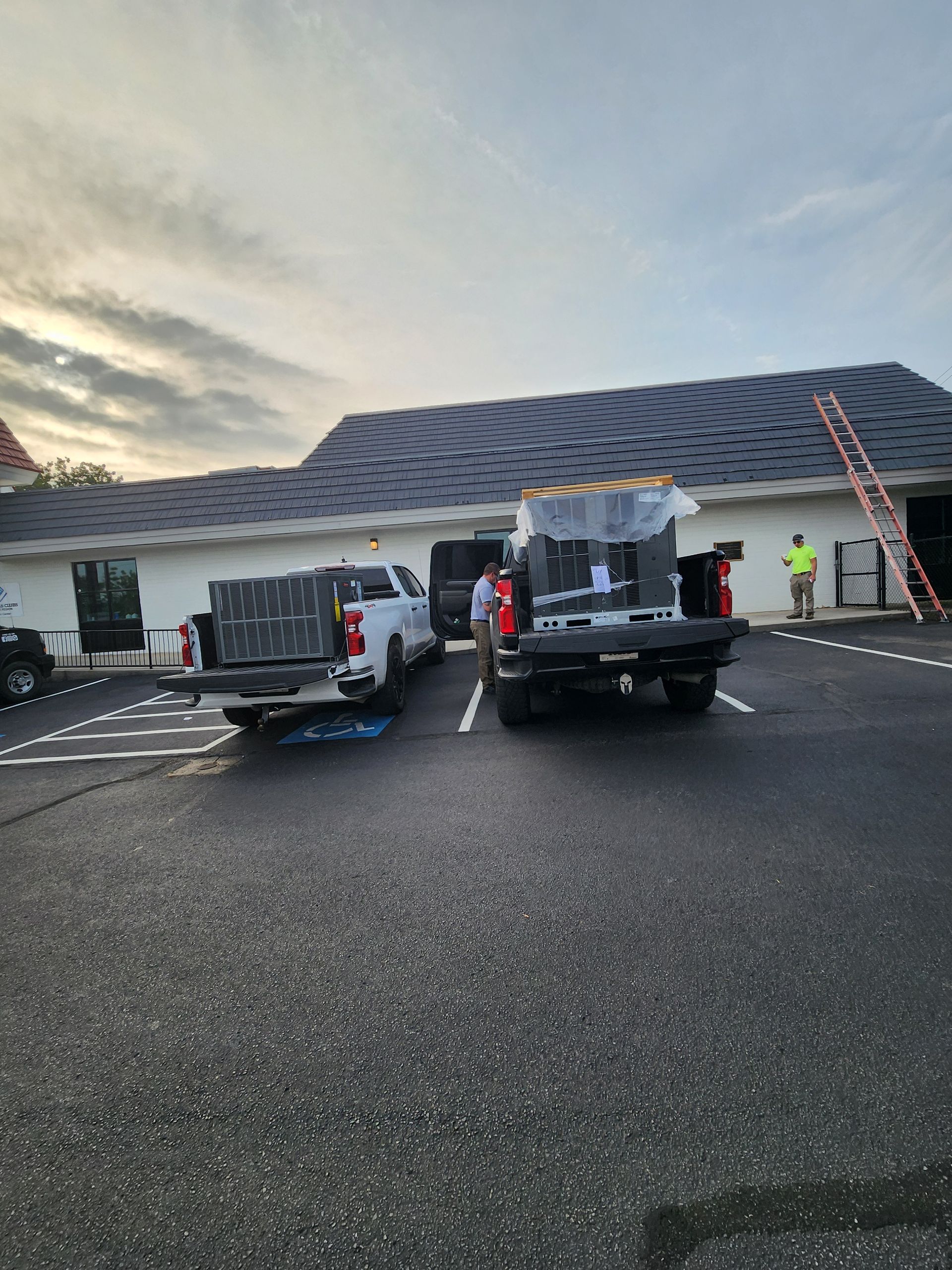 Construction vehicles and workers near a white building at dusk, with ladders and cones in a parking lot