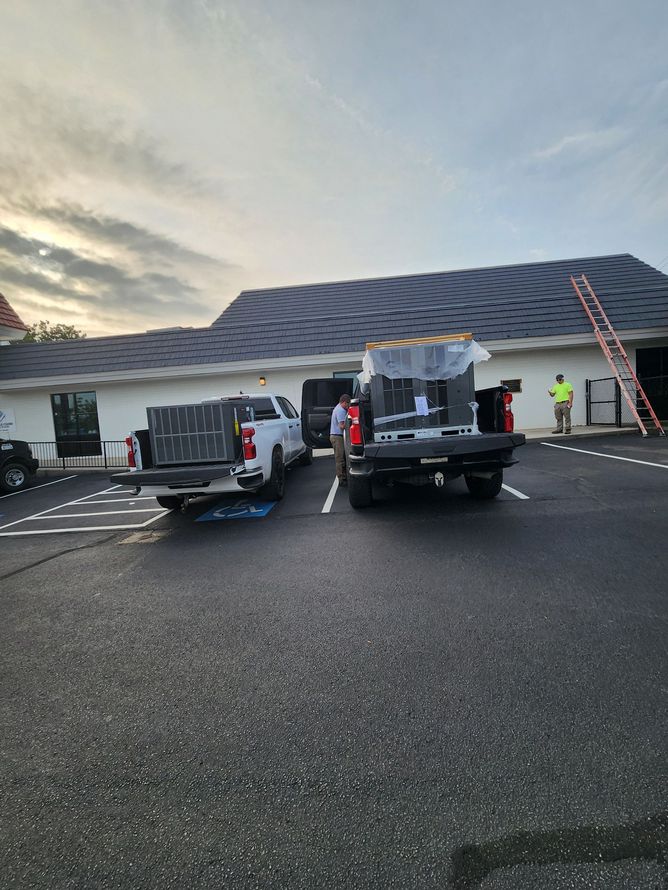 Construction vehicles and workers near a white building at dusk, with ladders and cones in a parking lot
