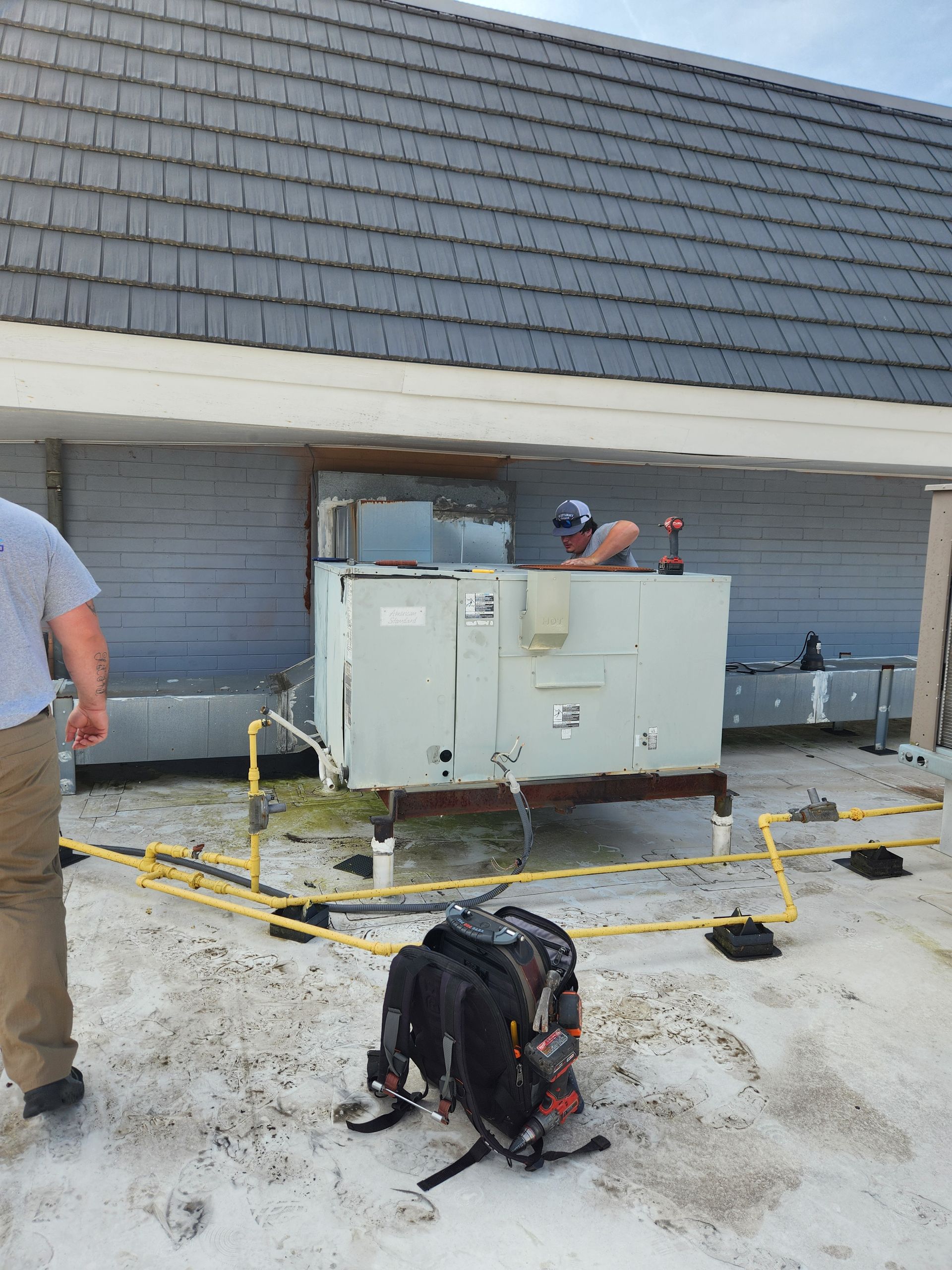 Roof repair workers beside an HVAC unit on a flat roof, with tools and a backpack in the foreground.