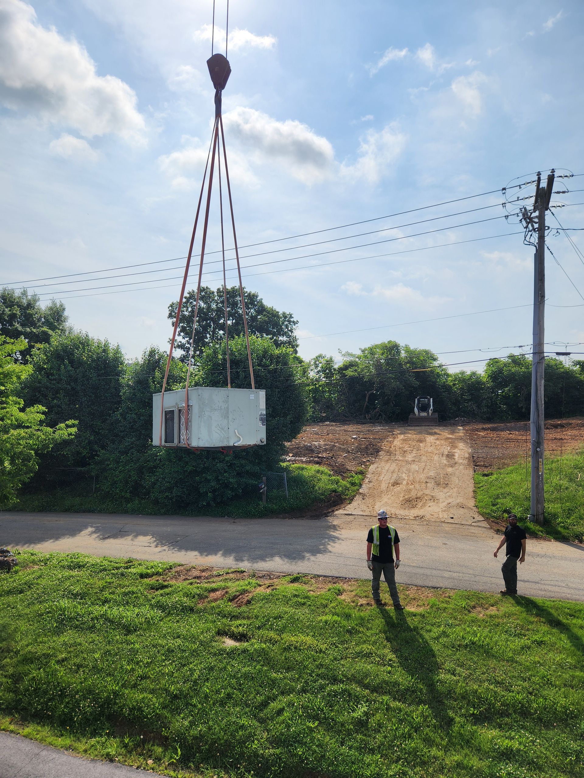 Two people near a white container suspended by red cables over a dirt road in a rural area