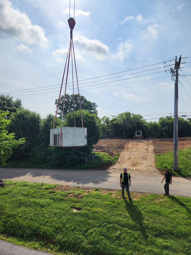Two people near a white container suspended by red cables over a dirt road in a rural area