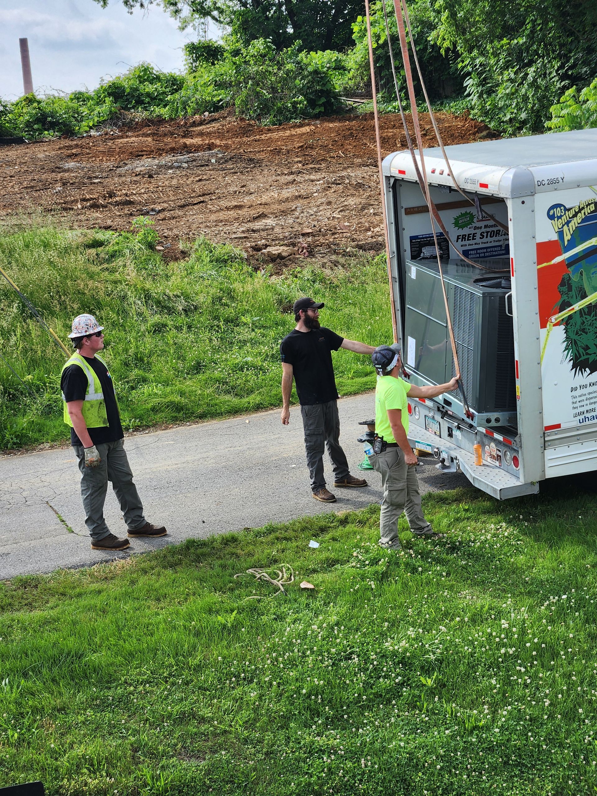 Workers unloading a white truck on a gravel path beside a grassy hill