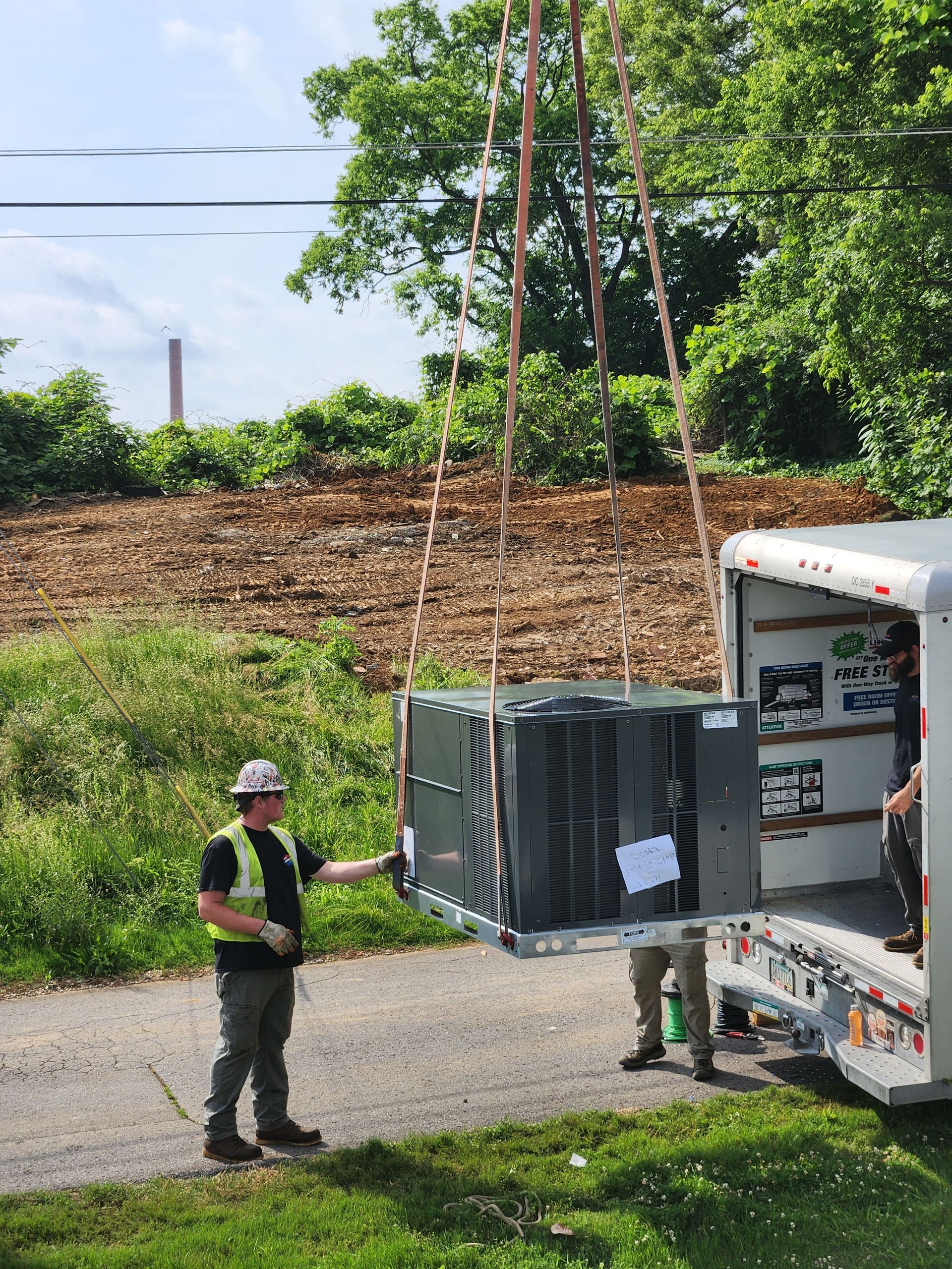 Two workers unloading a large HVAC unit from a truck on a roadside.