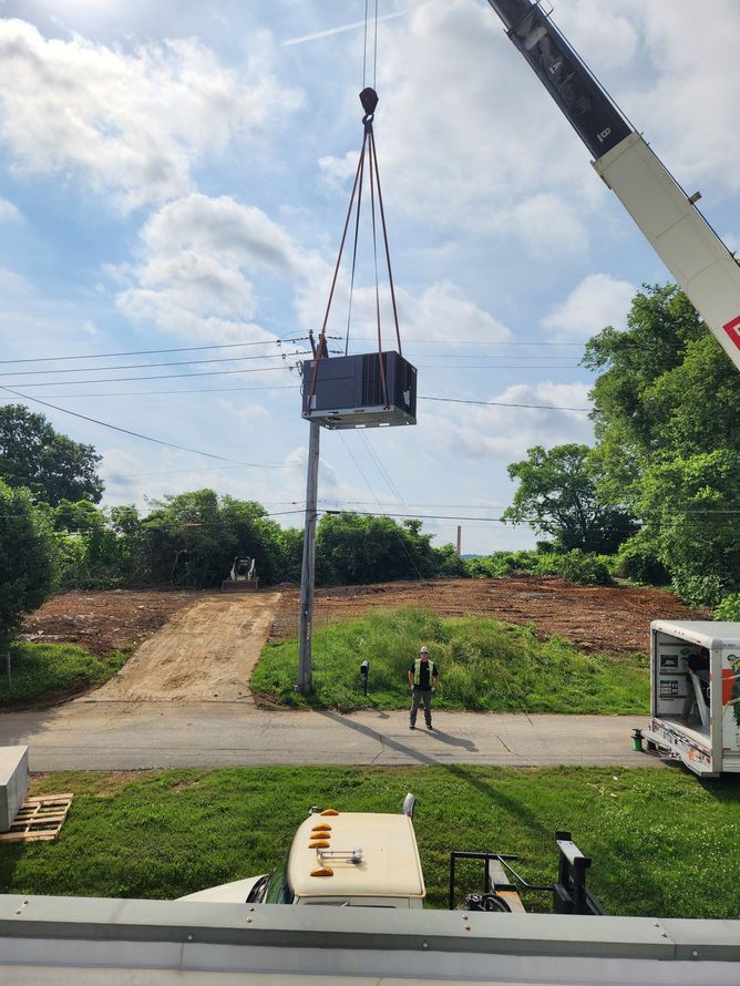 Crane lifting a dark box over a grassy outdoor site with trees and a small structure