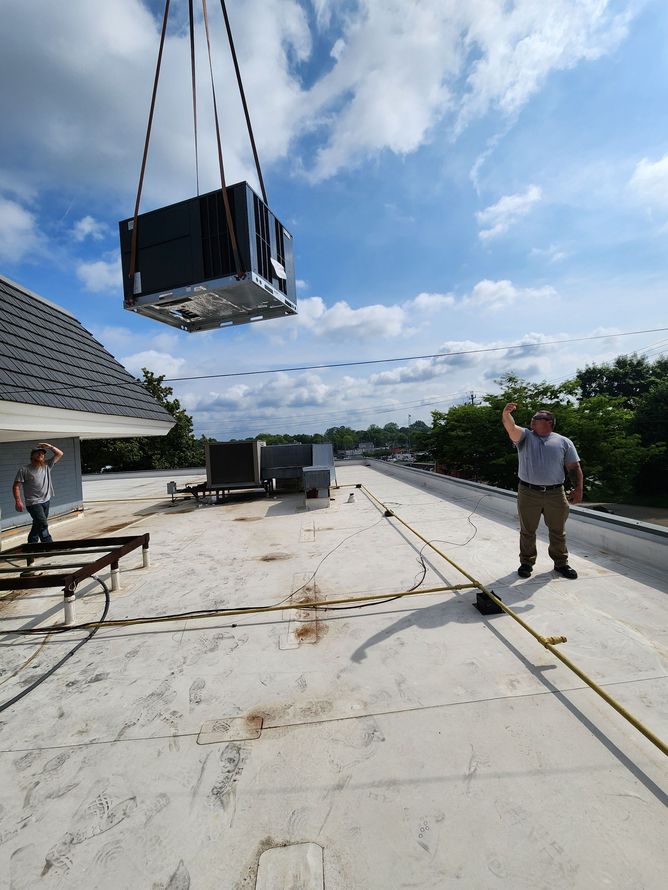 Roof workers lifting a black HVAC unit by crane on a flat rooftop under a cloudy sky