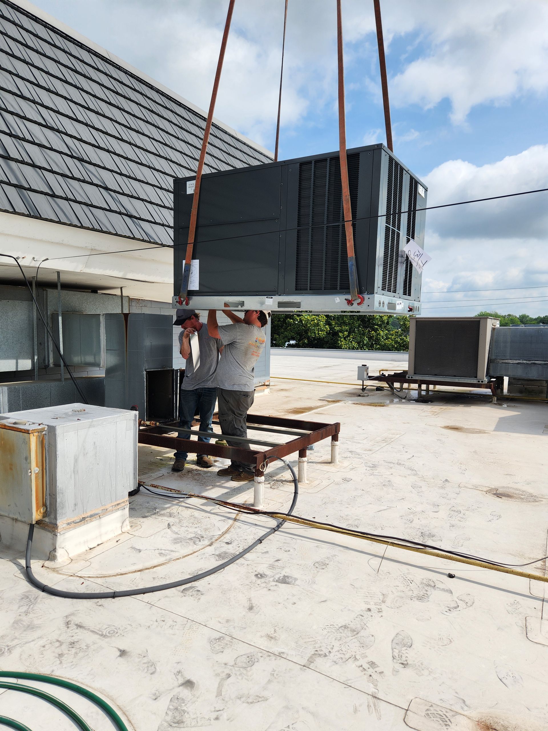 Worker hoisting a large black HVAC unit on a rooftop with straps and a crane.