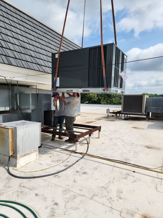 Worker hoisting a large black HVAC unit on a rooftop with straps and a crane.
