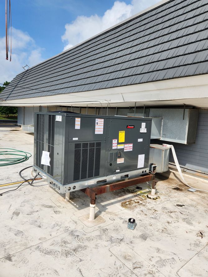 Industrial HVAC unit on a rooftop under a shingled roof, with pipes and wiring visible.