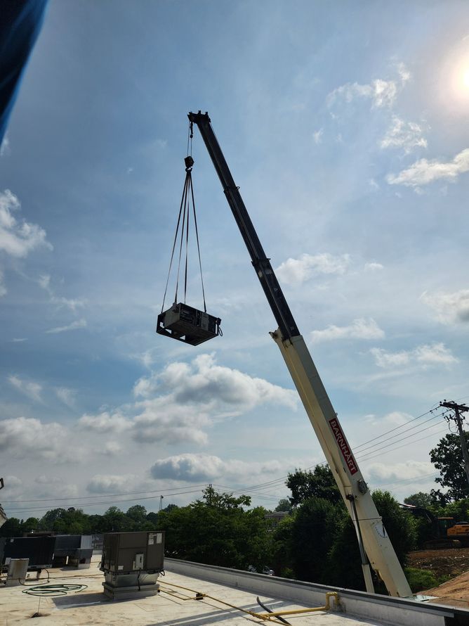 Crane lifting a large dark object high above a rooftop under a bright blue sky
