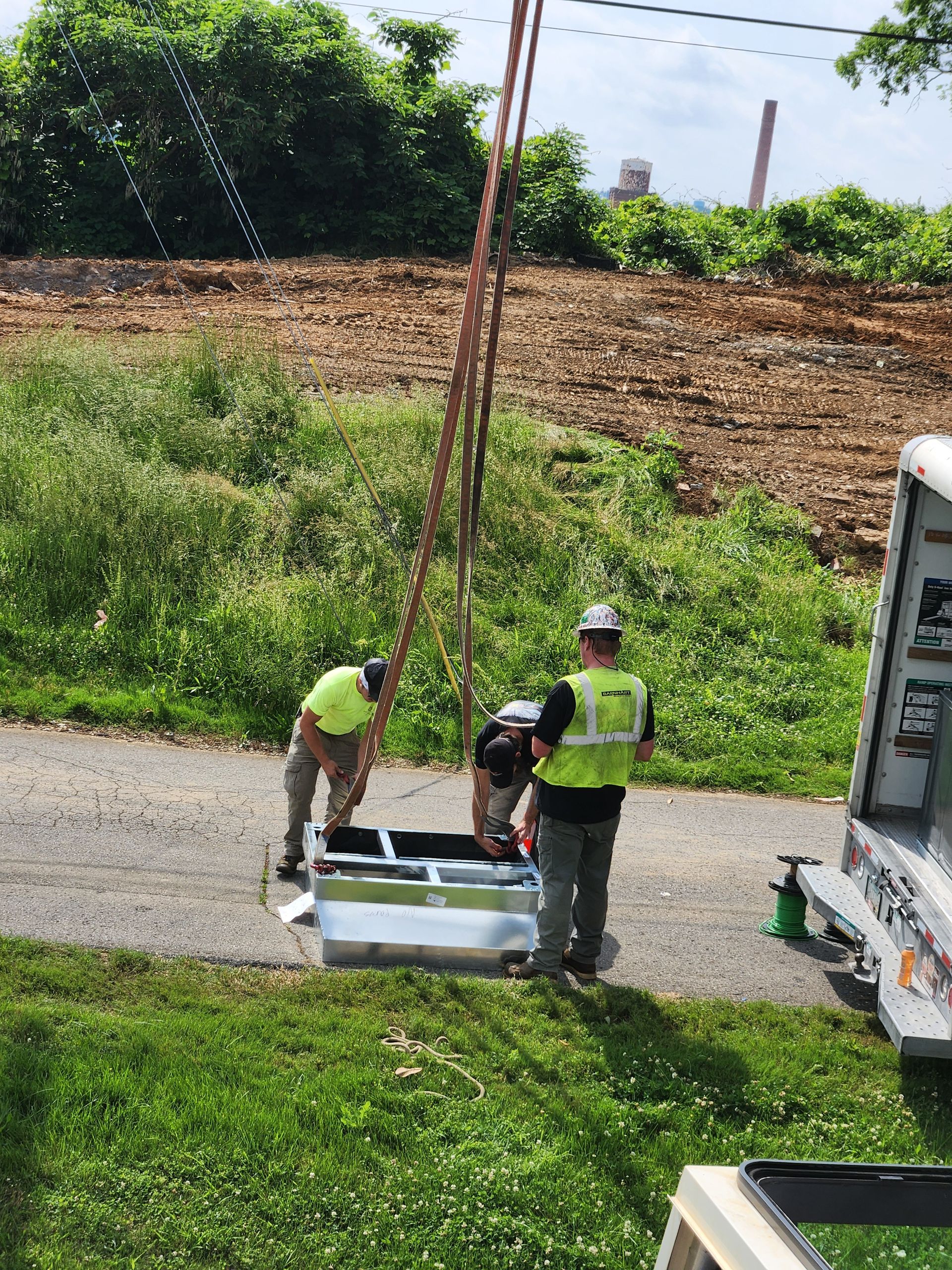 Workers in safety vests inspect a white utility box beside a road on a sunny day.