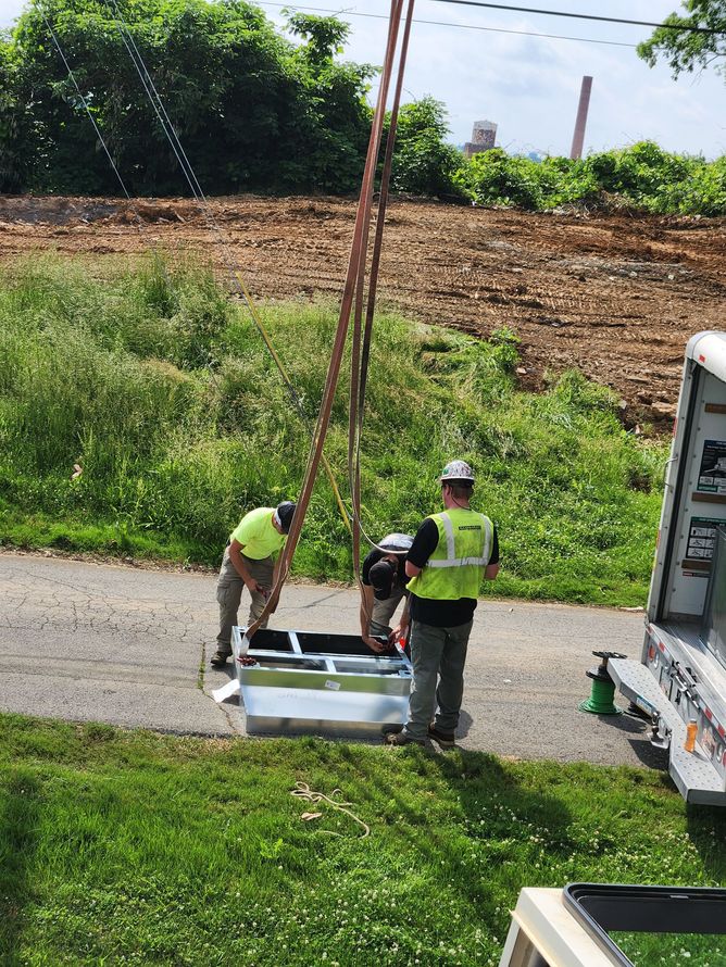 Workers in safety vests inspect a white utility box beside a road on a sunny day.