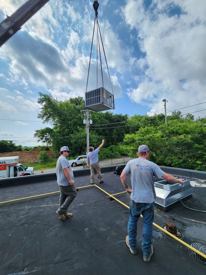 Workers using a crane to lift a large metal unit onto a roof under a cloudy sky