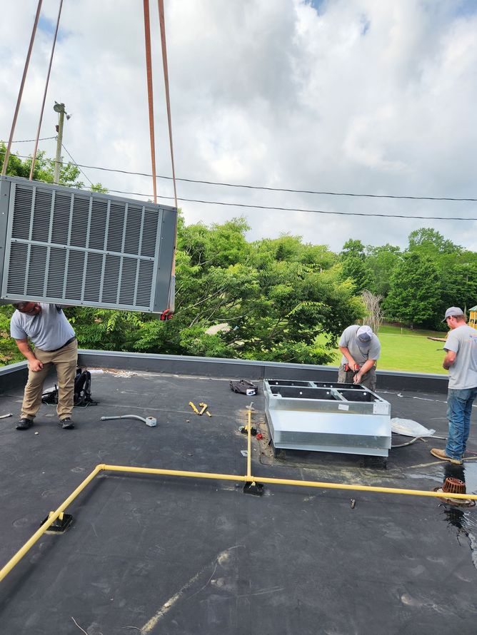 Workers on a rooftop lifting a large HVAC filter unit beside an open rooftop equipment panel.