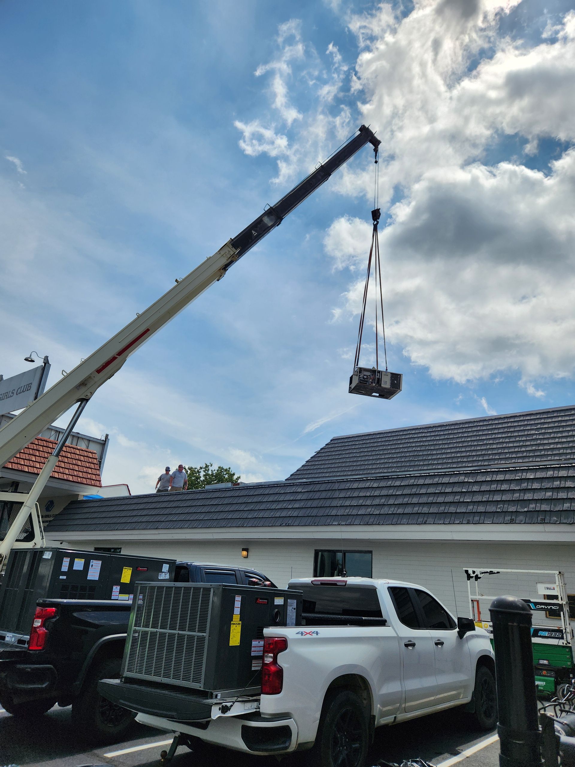 Crane lifting a load over a house roof, with white truck and workers below under a cloudy sky.