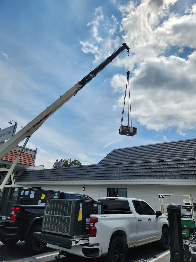 Crane lifting a load over a house roof, with white truck and workers below under a cloudy sky.