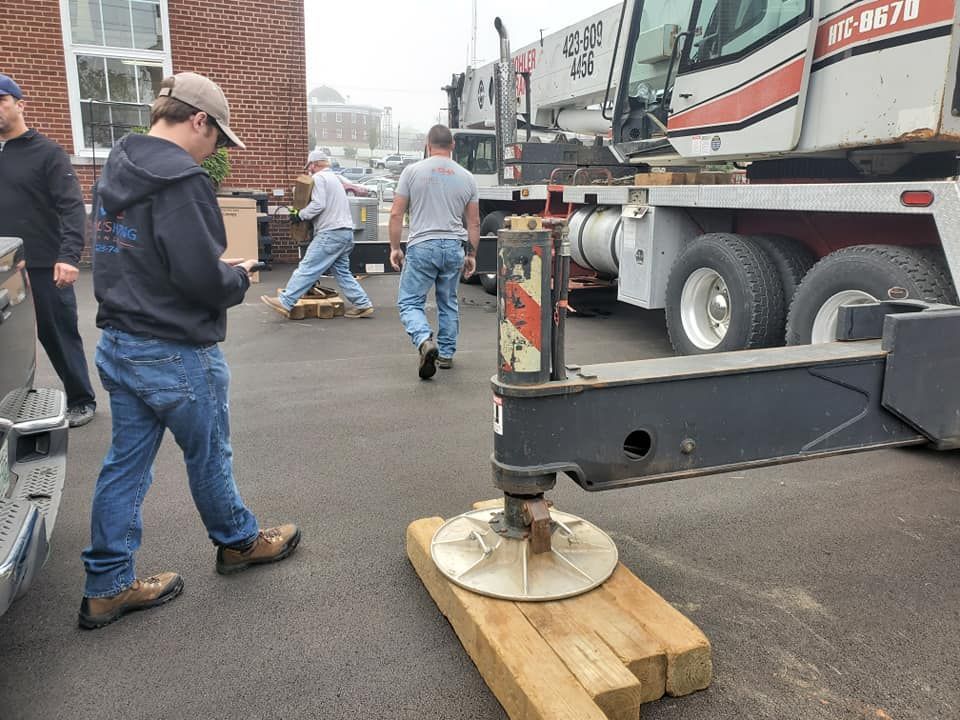 Workers moving equipment near a truck, with a metal base on wooden blocks in an outdoor work area