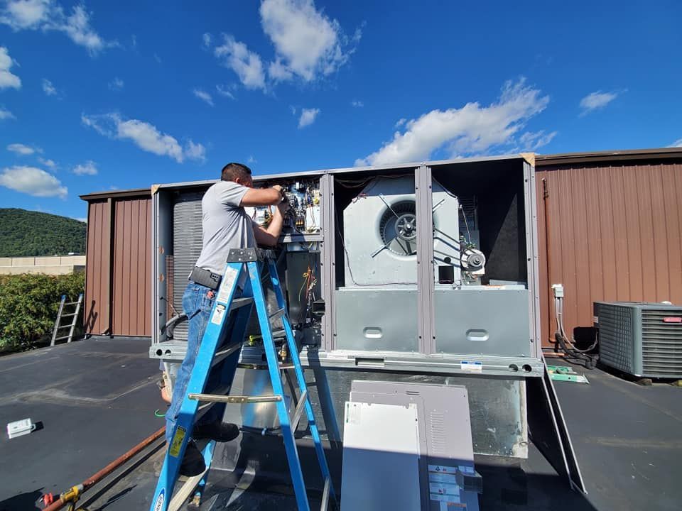 Worker on a blue ladder repairing HVAC equipment outdoors beside a brown building.