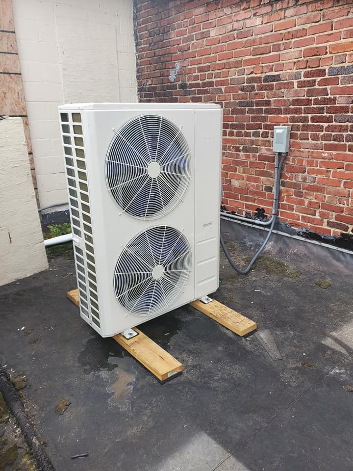 Outdoor HVAC condenser unit beside a brick wall on wooden blocks, with dual fans and a control box nearby