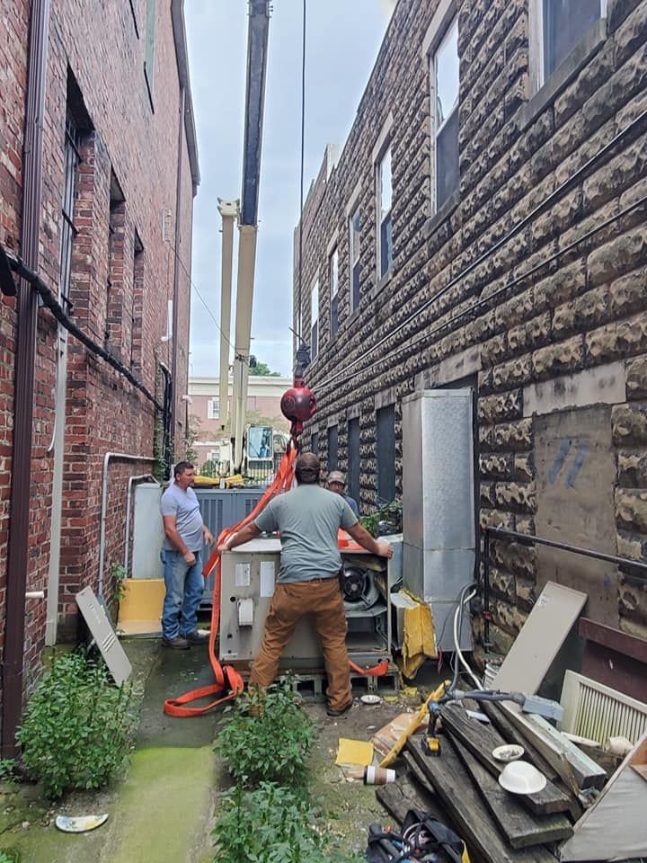 Two workers use an orange pump hose in a narrow alley between brick buildings, with tools and debris nearby.